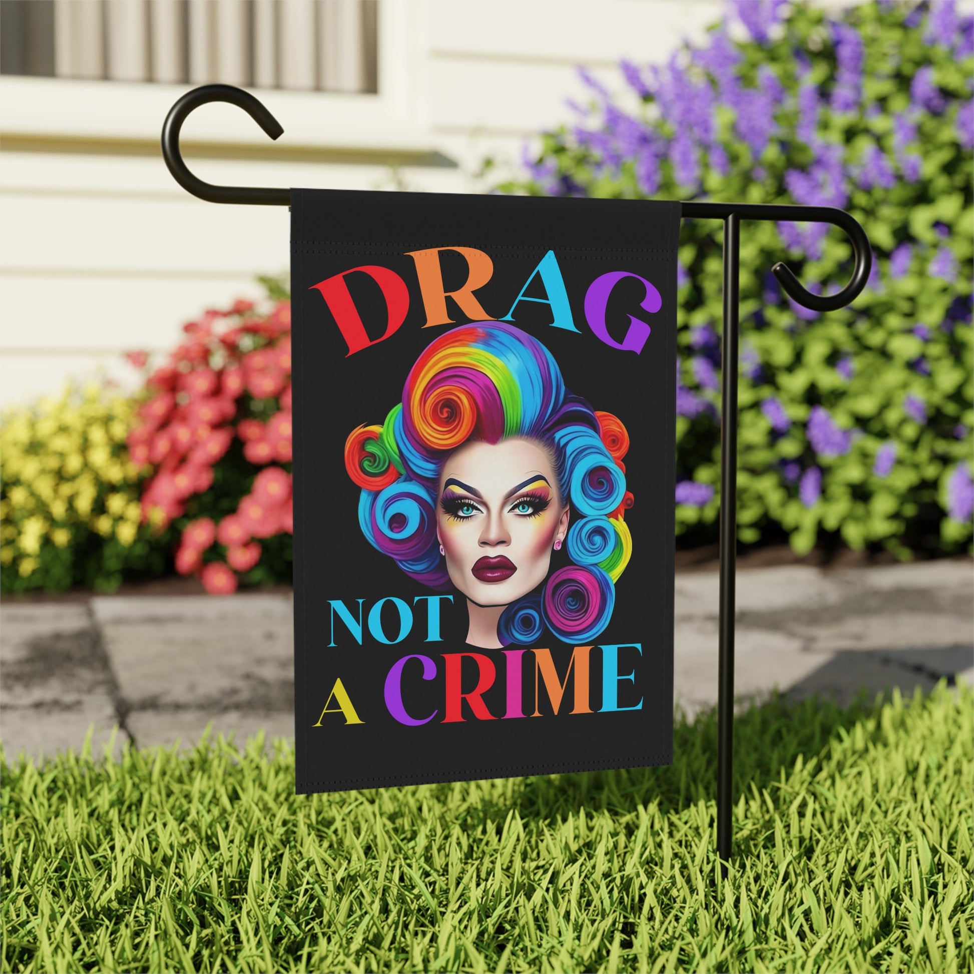 Garden flag with drag queen design and 'Drag Not A Crime' text in a colorful font, placed on grass with flowers in the background.