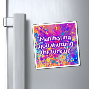 A square refrigerator magnet with a colorful paint splatter background and the text 'Manifesting you shutting the fuck up' in white with a purple outline, shown attached to a silver stainless steel refrigerator door next to the handle.