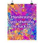 A colorful poster print with a vibrant paint splatter background and the text 'Manifesting you shutting the fuck up' in white letters with a purple outline, hanging from two black binder clips against a white background.