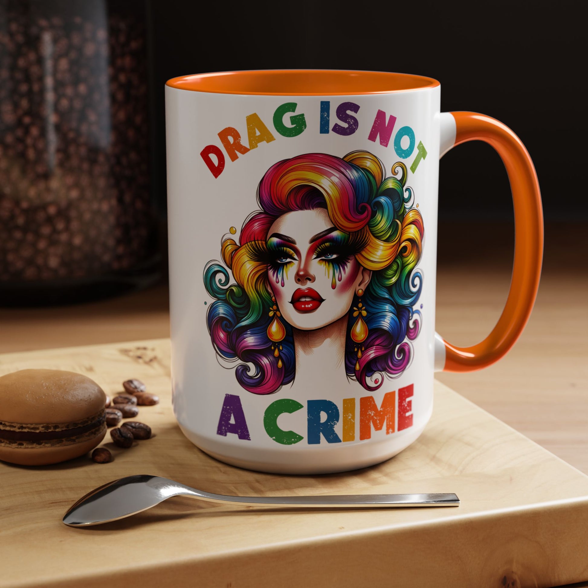Mug with colorful design and text 'DRAG IS NOT A CRIME' on a wooden surface with coffee beans and a macaron.