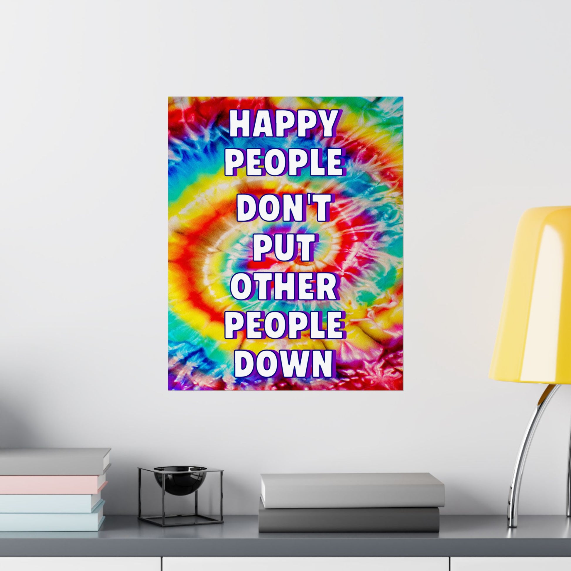 A square wall poster with a colorful tie-dye spiral background and the text 'HAPPY PEOPLE DON'T PUT OTHER PEOPLE DOWN' in bold white letters with a purple outline, displayed on a light gray wall above a shelf with books and a yellow lamp.