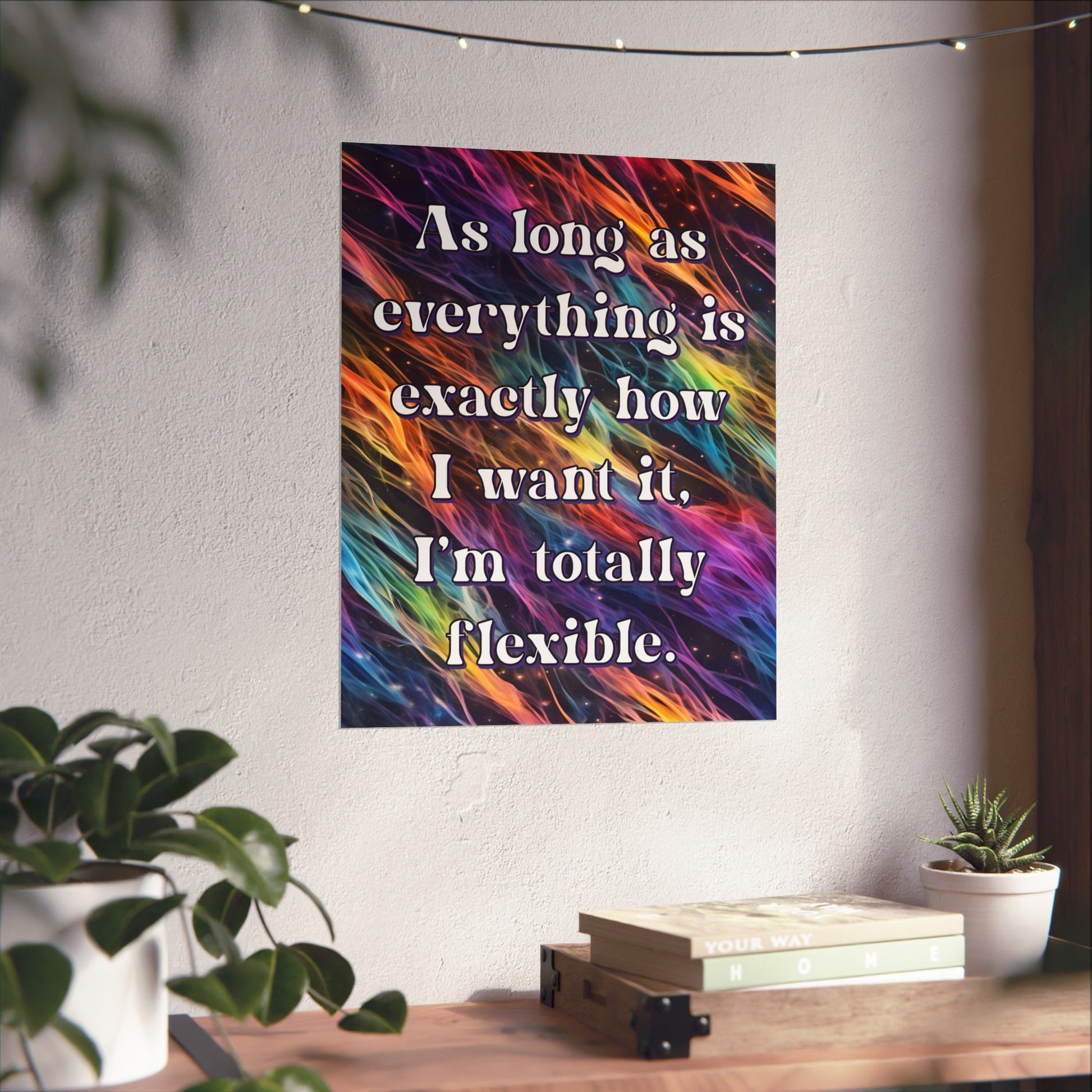 A vertical wall art print with a colorful abstract brushstroke background and the quote 'As long as everything is exactly how I want it, I'm totally flexible.' in white serif text, displayed on a light wall above a wooden surface with books and small plants.
