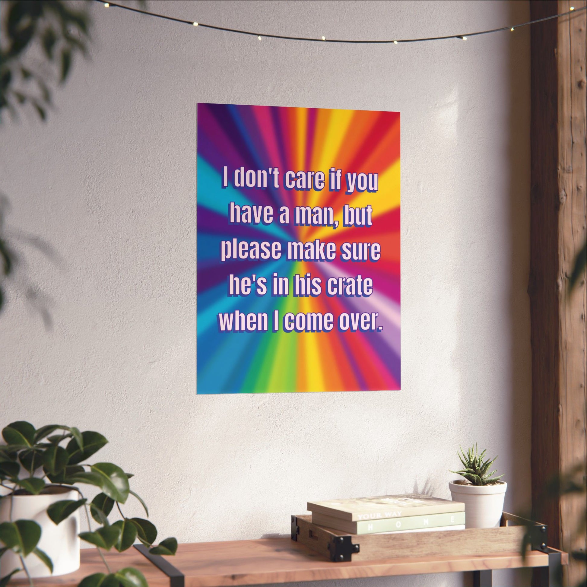 A rectangular wall poster with a rainbow burst background and white text reading, 'I don't care if you have a man, but please make sure he's in his crate when I come over,' displayed on a light wall above a wooden console table with plants.