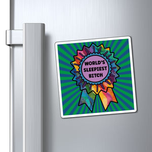 A square refrigerator magnet with a colorful rainbow ribbon award design and the text 'WORLD'S SLEEPIEST BITCH' on a green sunburst background, shown attached to a silver refrigerator door.