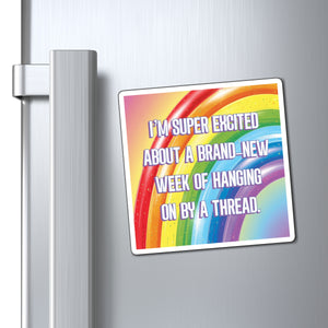 A square refrigerator magnet with a colorful rainbow design and white text reading, 'I'M SUPER EXCITED ABOUT A BRAND NEW WEEK OF HANGING ON BY A THREAD,' shown attached to a silver refrigerator door next to the handle.
